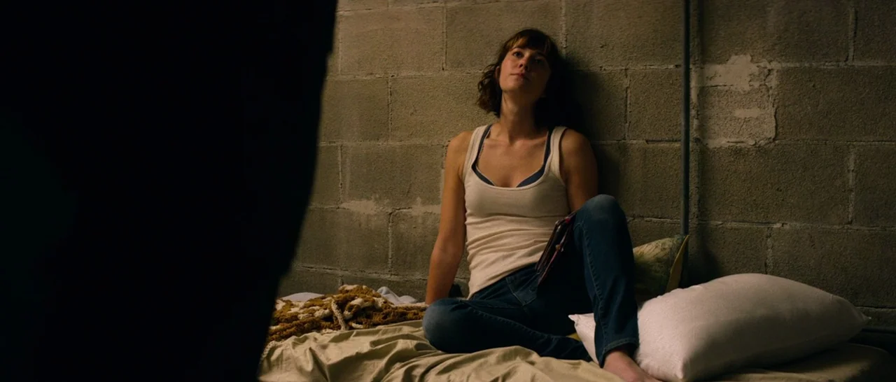 10 Cloverfield Lane (2016) movie still: medium — A dark, cramped space with unfinished cinder block walls and a simple sleeping area. A woman in a…