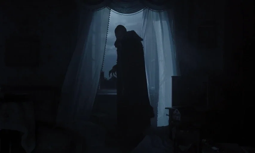 Nosferatu (2024) movie still: fullbody — A dimly lit room with sheer white curtains hanging in front of a window. A tall, cloaked silhouette of a…
