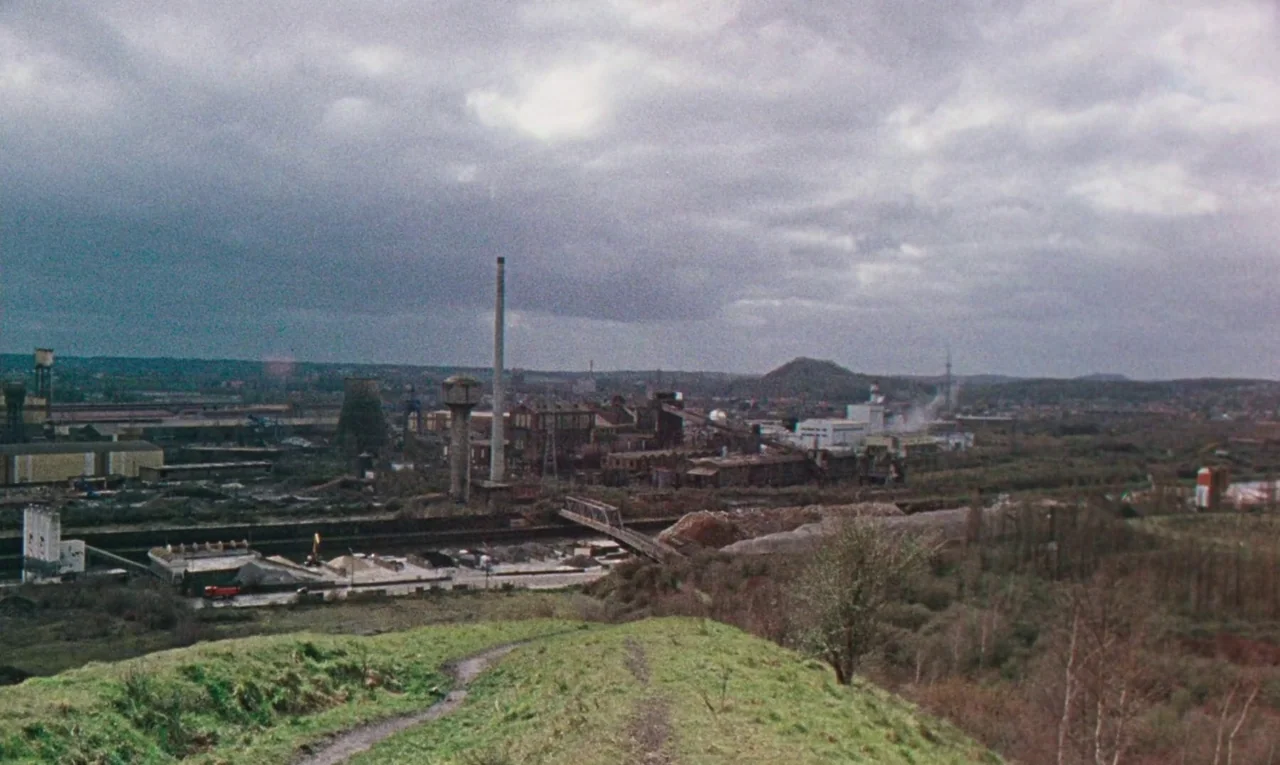 Maldoror (2024) movie still: establishing — An industrial manufacturing plant sits in a valley under a heavy, overcast cloudy sky. Foreground grass…