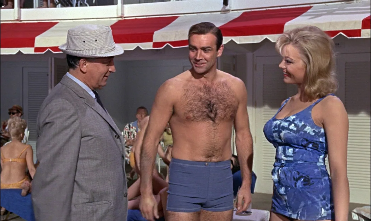 Goldfinger (1964) movie still: medium — A sunlit pool deck features a red and white striped overhead awning. Three individuals are positioned in the…
