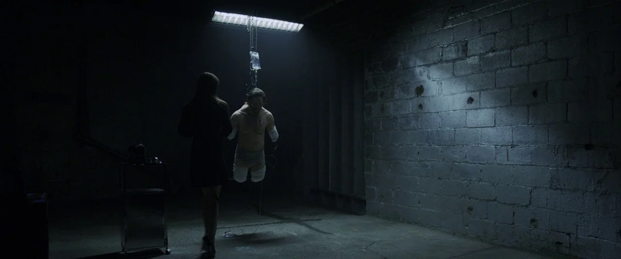 American Mary (2012) movie still: A cold, clinical interrogation or torture scene set in an industrial bunker. The harsh top-down lighting…