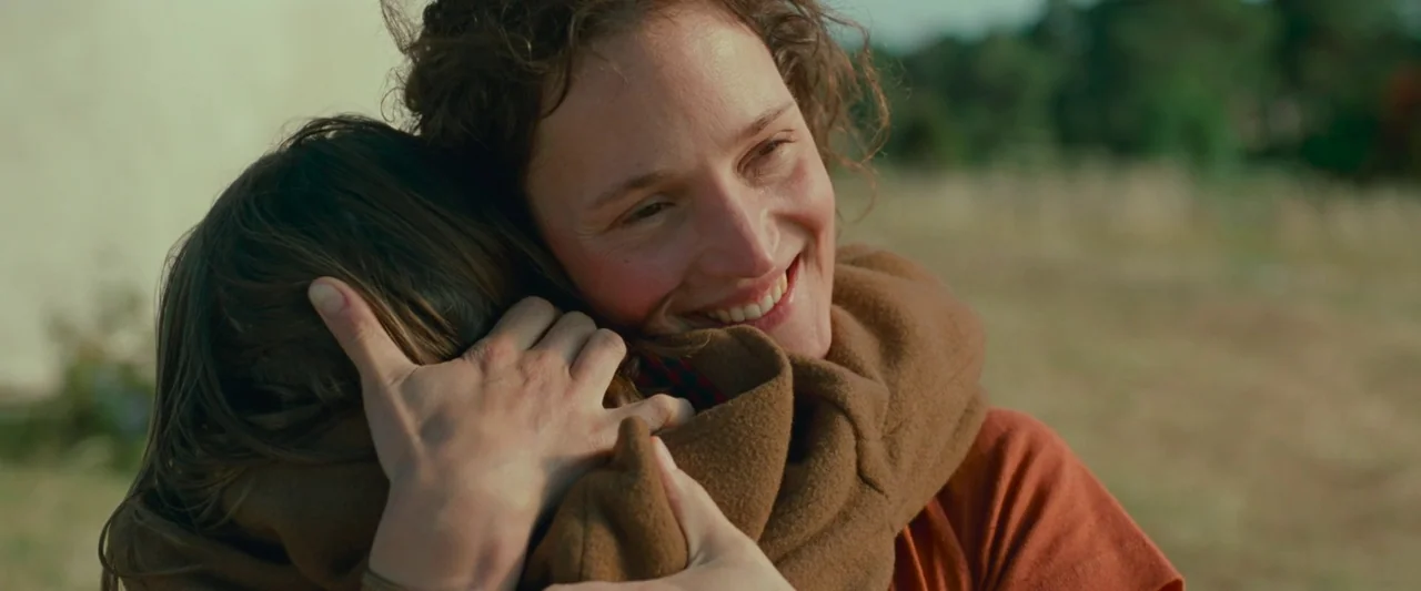 Bergman Island (2021) movie still: A close-up shot capturing a tender, intimate hug between an adult and a child in an outdoor setting. The warm…