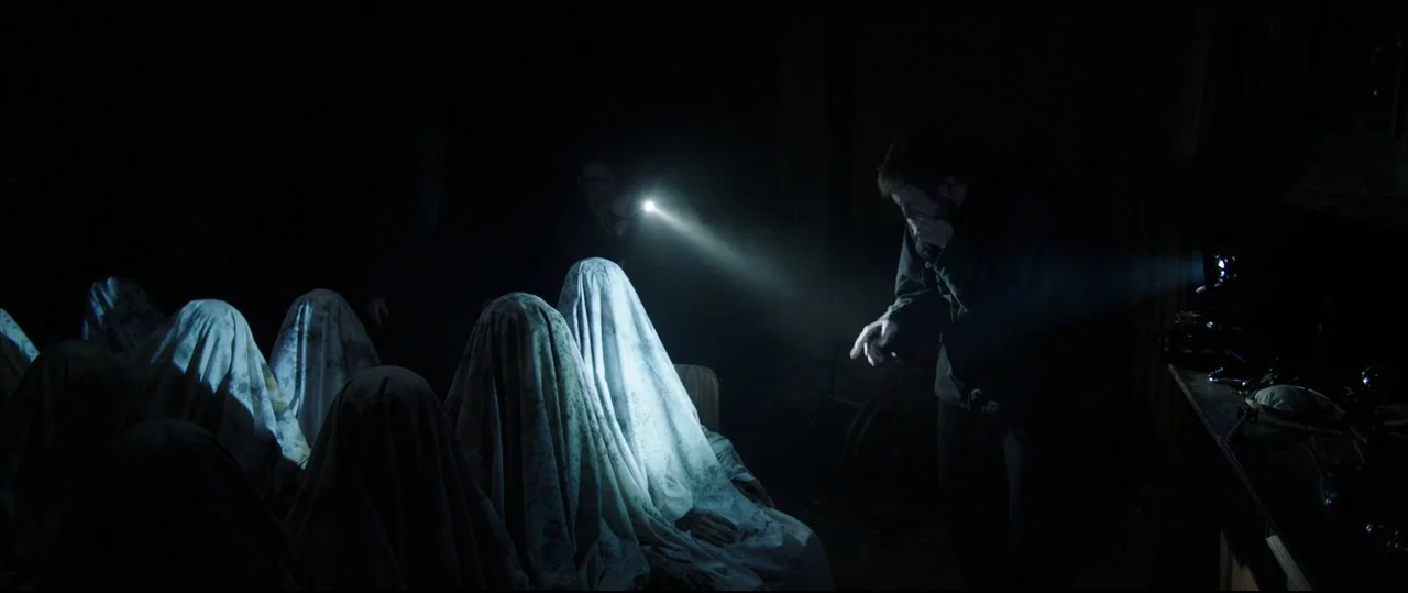 Insidious: Chapter 2 (2013) movie still: medium — A dark, cluttered space features multiple figures seated in a row covered by white sheets. A man…