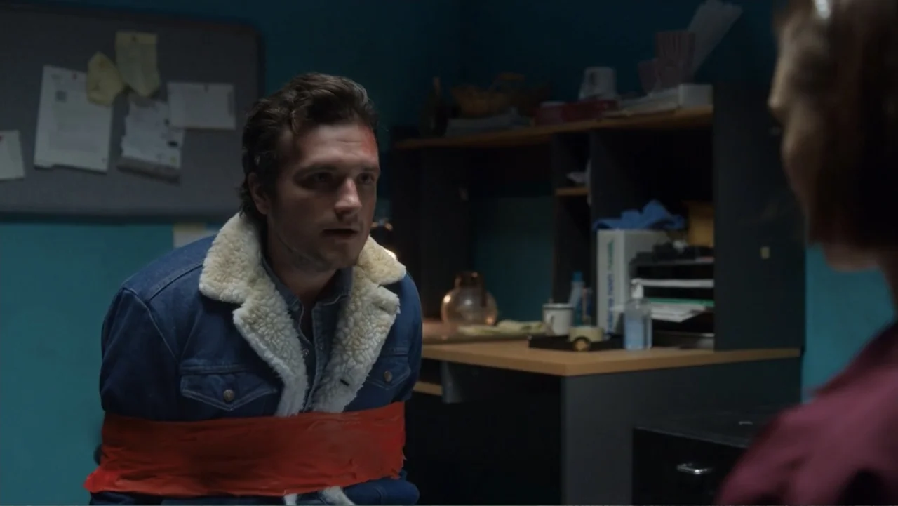 Burn (2019) movie still: medium — An office space with teal walls and a cluttered desk containing a lamp. A man sits in a chair restrained with red…