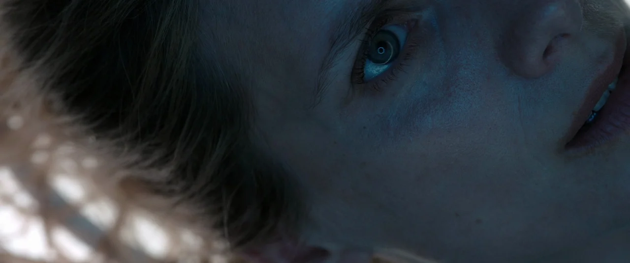 Oxygen (2021) movie still: closeup — A close-up view of a woman's face illuminated by a cold light source. Her eye reflects a circular light pattern…