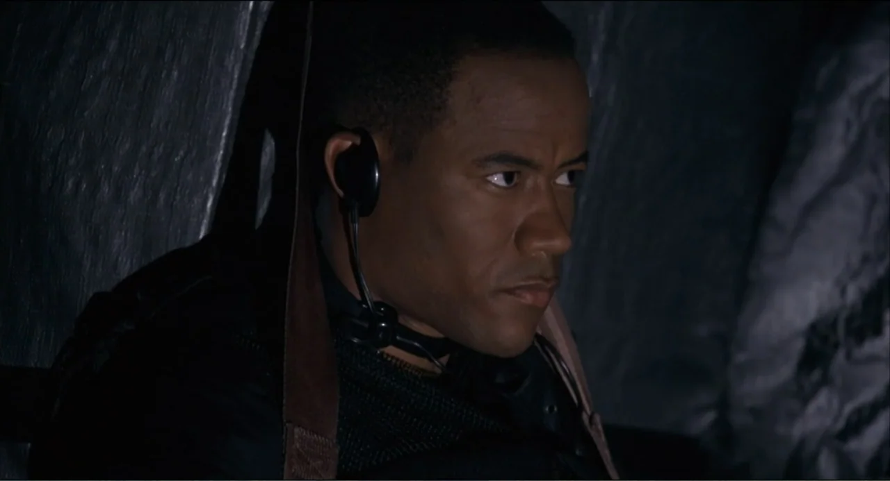 Air Rage (2001) movie still: closeup — A dark, cramped interior of an aircraft cockpit with metallic surfaces. A man wearing a black headset looks…