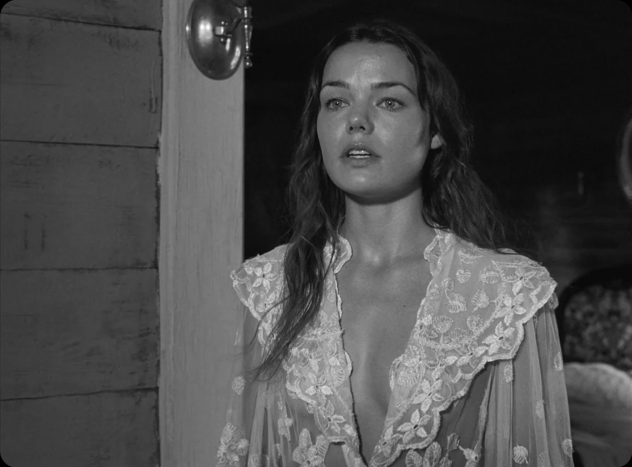 Eureka (2024) movie still: medium — A dimly lit room with wooden wall paneling and a wall sconce. A woman wears a delicate white lace garment while…