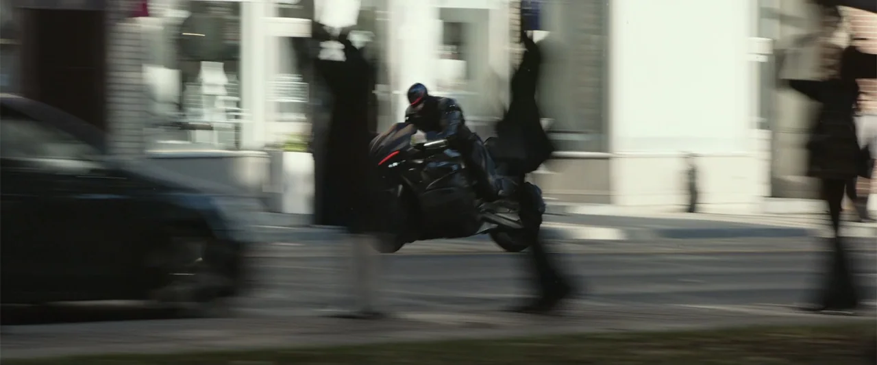 RoboCop (1987) movie still: medium — A blur of motion captures a motorcyclist speeding through a bright urban street intersection. The rider leans…