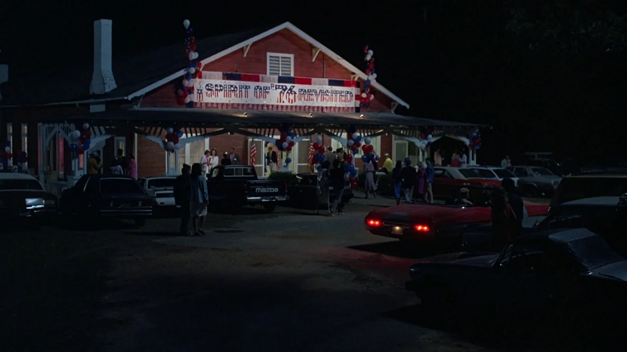 Something Wild (1986) movie still: wide — A wooden community building is decorated with red, white, and blue balloons and banners. People walk among…