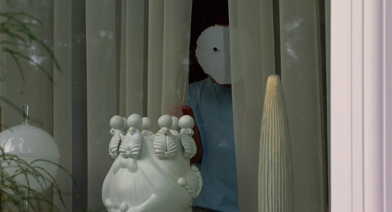 Hatching (2022) movie still: medium — A light-filled interior space contains decorative white sculptures on a surface behind a window. A person…