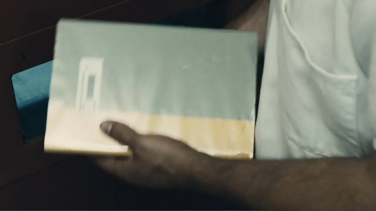 Camp X-Ray (2014) movie still: closeup — An interior workspace with a person holding a book near a partition wall. The person holds a light green…