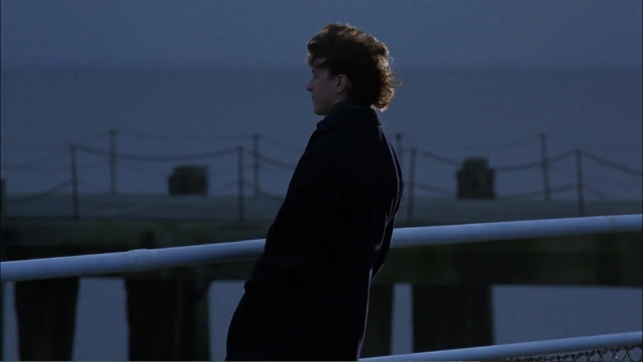 Starter for 10 (2006) movie still: medium — A wooden pier extends into calm dark water during the blue hour at twilight. A man in a dark coat stands…