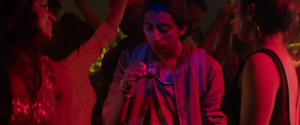 Dope (2015) movie still: medium — A dimly lit nightclub space filled with silhouettes of dancing people. A man stands centrally holding a small…