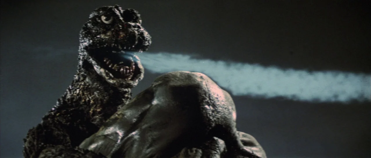 Godzilla Vs Hedorah (1971) movie still: medium — A dark, nondescript night environment with a textured monster and a foreground object. A large…