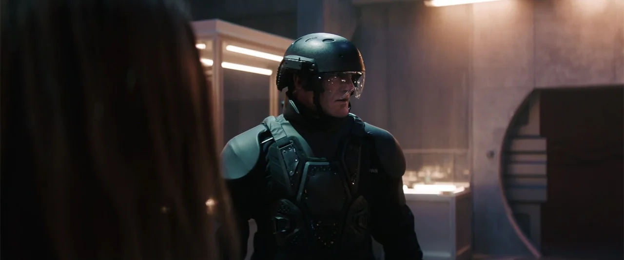 M3GAN 2.0 (2025) movie still: medium — A dark industrial corridor features concrete walls and illuminated cabinets in the background. A man wearing…