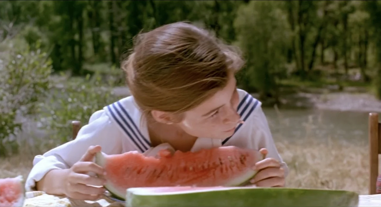 A River Runs Through It (1992) movie still: medium — A young woman sits at a table outdoors near a grassy riverbank. She holds a large slice of…
