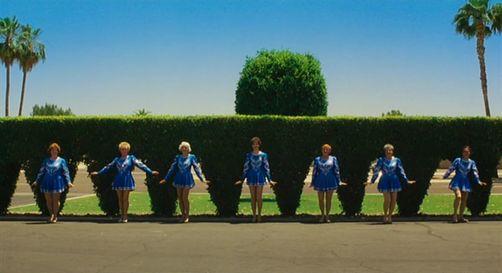 The Savages (2007) movie still: wide — Seven women stand in a row before a precisely sculpted, inverted-triangular green hedge. The paved surface is…