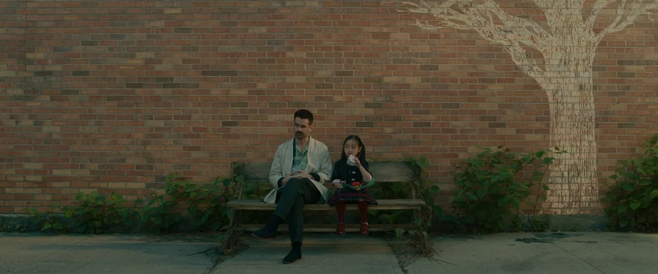 After Yang (2021) movie still: wide — A man and a young girl sit on a rustic bench against a brick wall. A large white tree mural is painted on the…