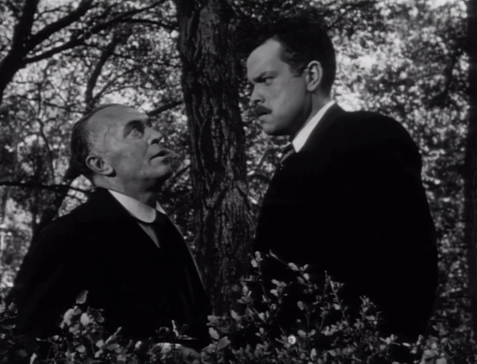 The Stranger (1946) movie still: medium — Two men converse in a dense outdoor wooded area with trees surrounding them. One man stands taller than…