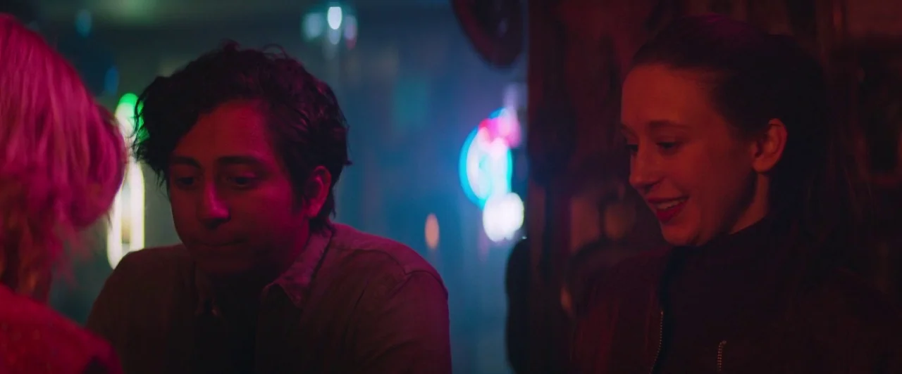 The Long Dumb Road (2018) movie still: medium — A dimly lit tavern with vibrant neon signs glowing in the blurry background. A young man and woman…