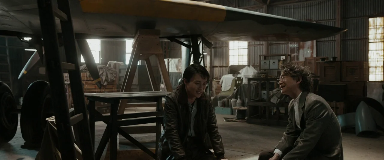 Godzilla Minus One (2023) movie still: medium — An industrial hangar features corrugated metal walls and a large, vintage airplane under…