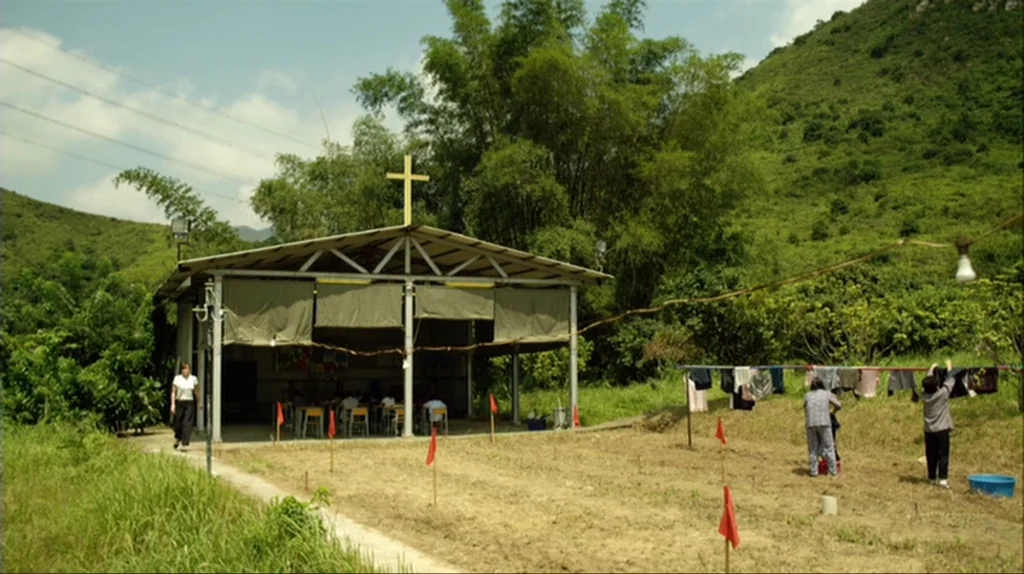 Contagion (2011) movie still: wide — A rural setting features a small pavilion with a cross topped roof among bamboo. People are occupied with…