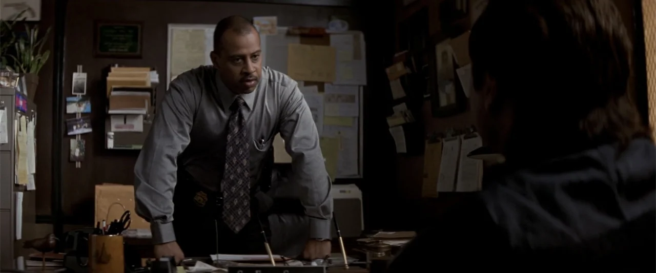 Angst (2017) movie still: medium — A cluttered police office features dark wood paneling, a corkboard, and paper files. An officer in a dress shirt…