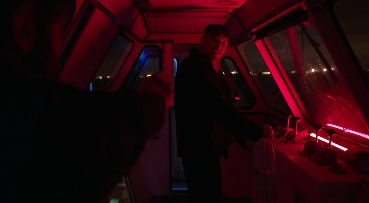 Urge (2016) movie still: medium — A dimly lit boat pilothouse at night illuminated by harsh red neon light. A man operates the steering wheel while…