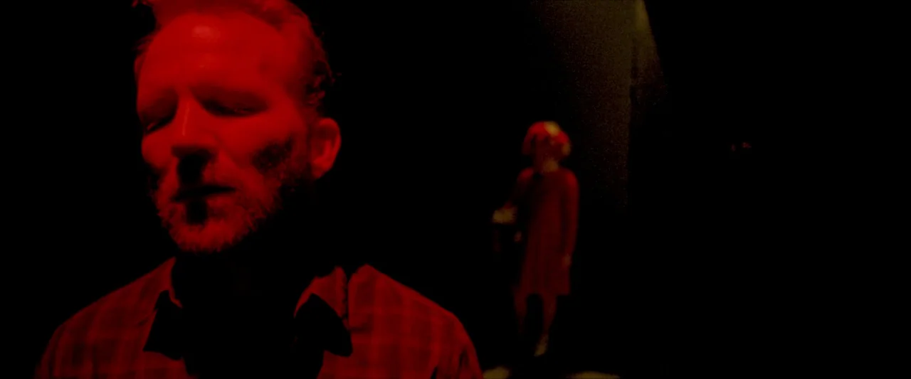 Moon Garden (2022) movie still: closeup — A dimly lit room is filled with intense red light against solid black shadows. A man with a beard stands…