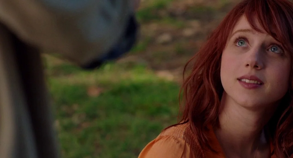 Ruby Sparks (2012) movie still: closeup — A woman stands in a green park filled with grass. She looks upward at an unseen person positioned in the…
