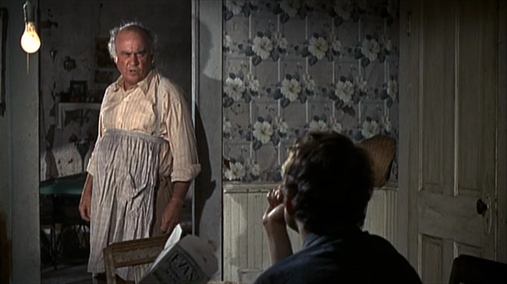Bonnie And Clyde (1967) movie still: medium — A dimly lit room features floral wallpaper and a wooden door on the right. An elderly man wearing an…