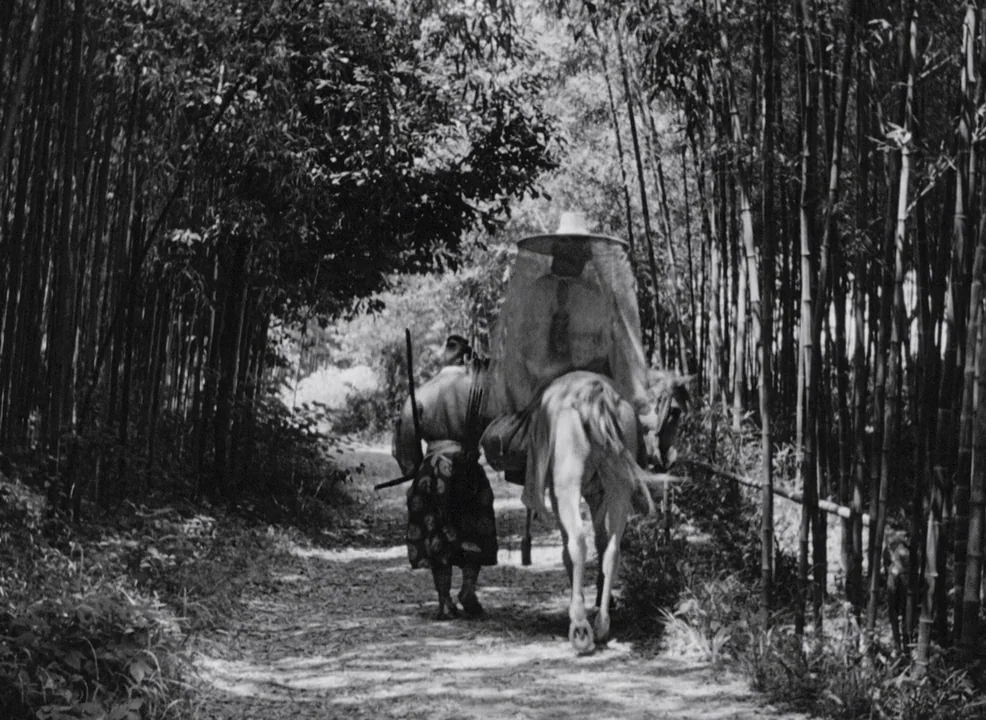 Rashomon (1950) movie still: fullbody — A dirt path cuts through a dense thicket of tall slender bamboo trees. A rider on a horse and a companion on…