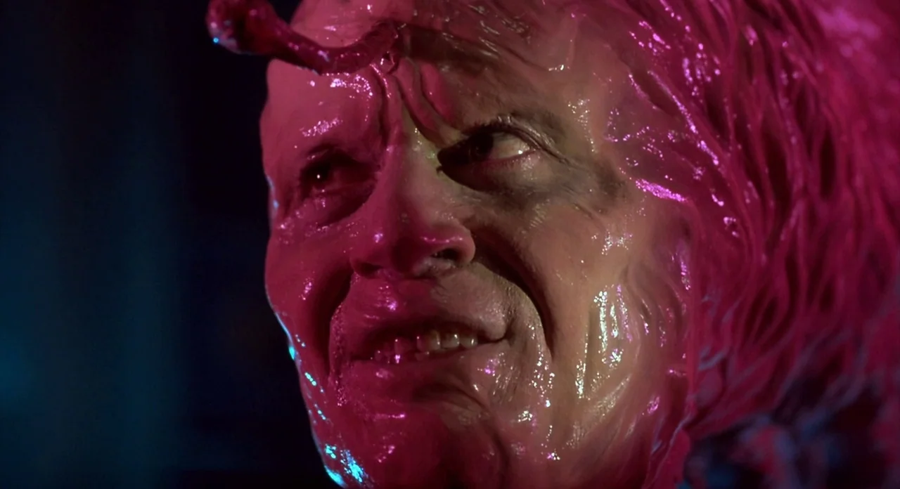 From Beyond (1986) movie still: closeup — A man's face is covered in wet, fleshy, pulsating growths under intense colored light. He looks away from…