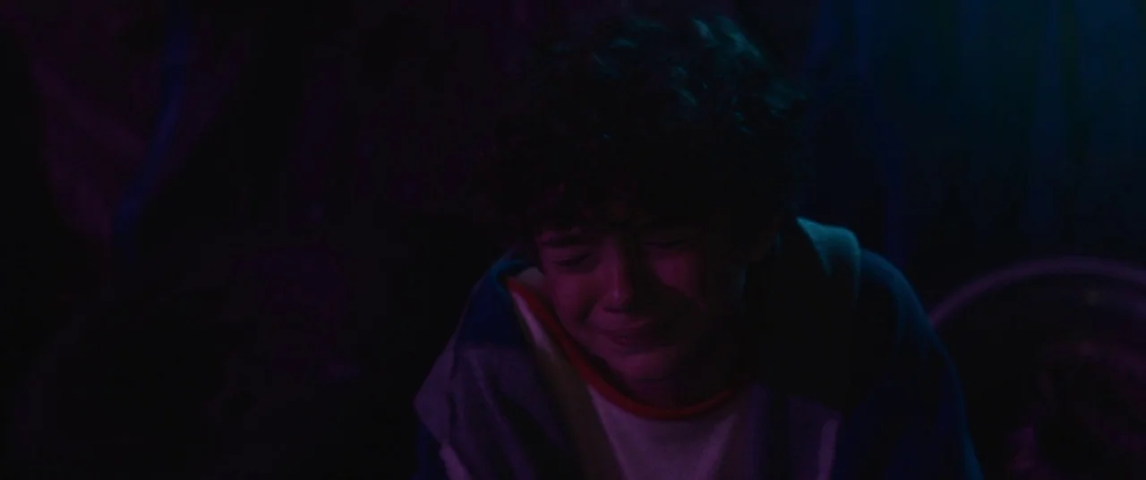 Honey Boy (2019) movie still: closeup — A dark, dimly lit room is filled with purple light and deep shadows. A young boy sits in the shadows with…