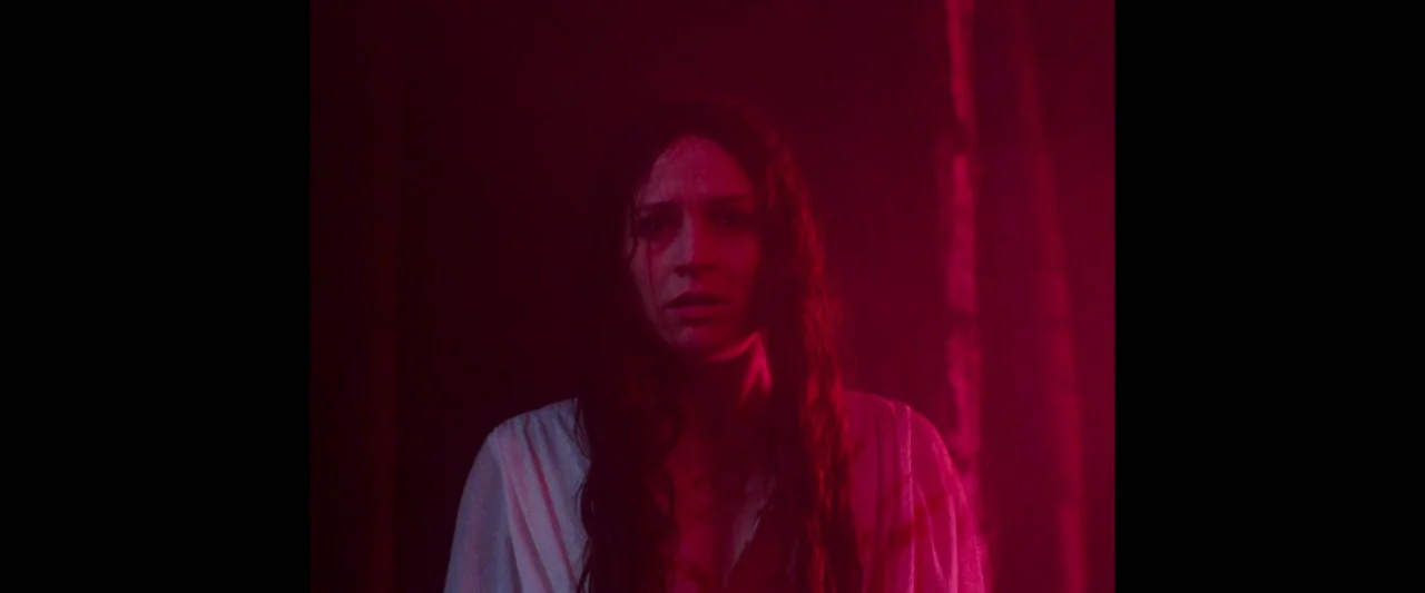 Censor (2021) movie still: medium — A woman stands within a dark area illuminated by a vibrant red light. She looks toward the camera while wearing…
