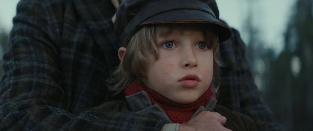 The Dead Don’t Hurt (2023) movie still: closeup — A child wearing a cap and red sweater is held by an adult outdoors. The adult's hand in a…