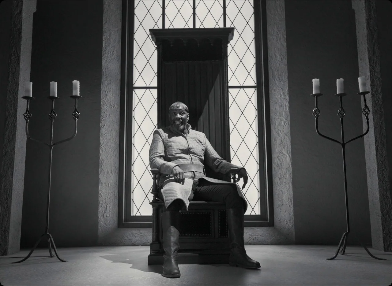 The Tragedy of Macbeth (2021) movie still: fullbody — A stone chamber features a large window with leaded glass behind a dark throne. A man sits…