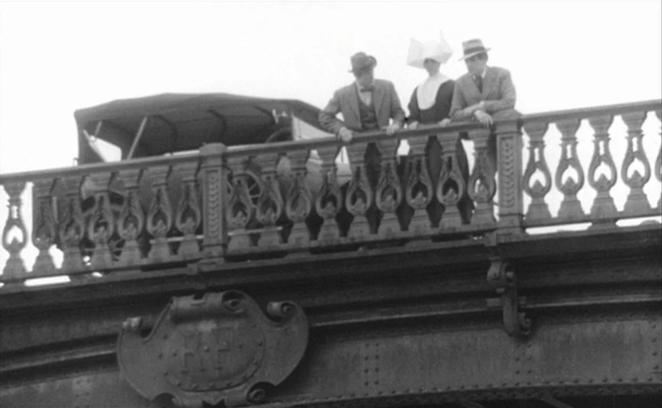 Judex (1966) movie still: fullbody — A metal bridge with ornate railings spans over a crossing. Two men and a nun in traditional habit lean against…