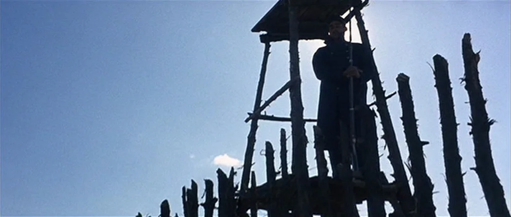 The Good, The Bad And The Ugly (1966) movie still: fullbody — A wooden watchtower stands against a bright, clear blue sky. A man in a dark coat…