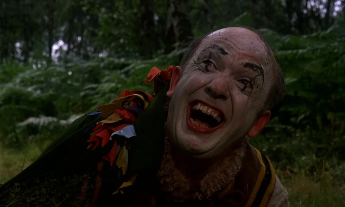 Vampire Circus (1972) movie still: closeup — A man with greasepaint makeup is surrounded by dense green ferns in a woodland. He looks toward the…