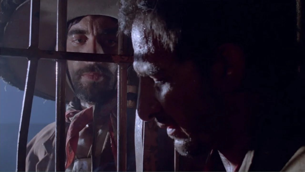 Toña Machetes (1985) movie still: medium — A dimly lit prison cell with vertical metal bars separates two male subjects. The men stand in close…