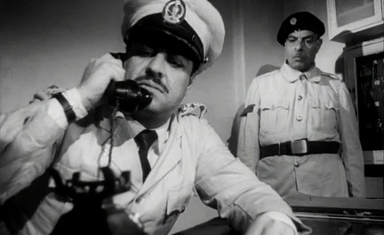 Cairo Station (1958) movie still: medium — A dimly lit room with plain walls features two men in military-style uniforms. A foreground officer…
