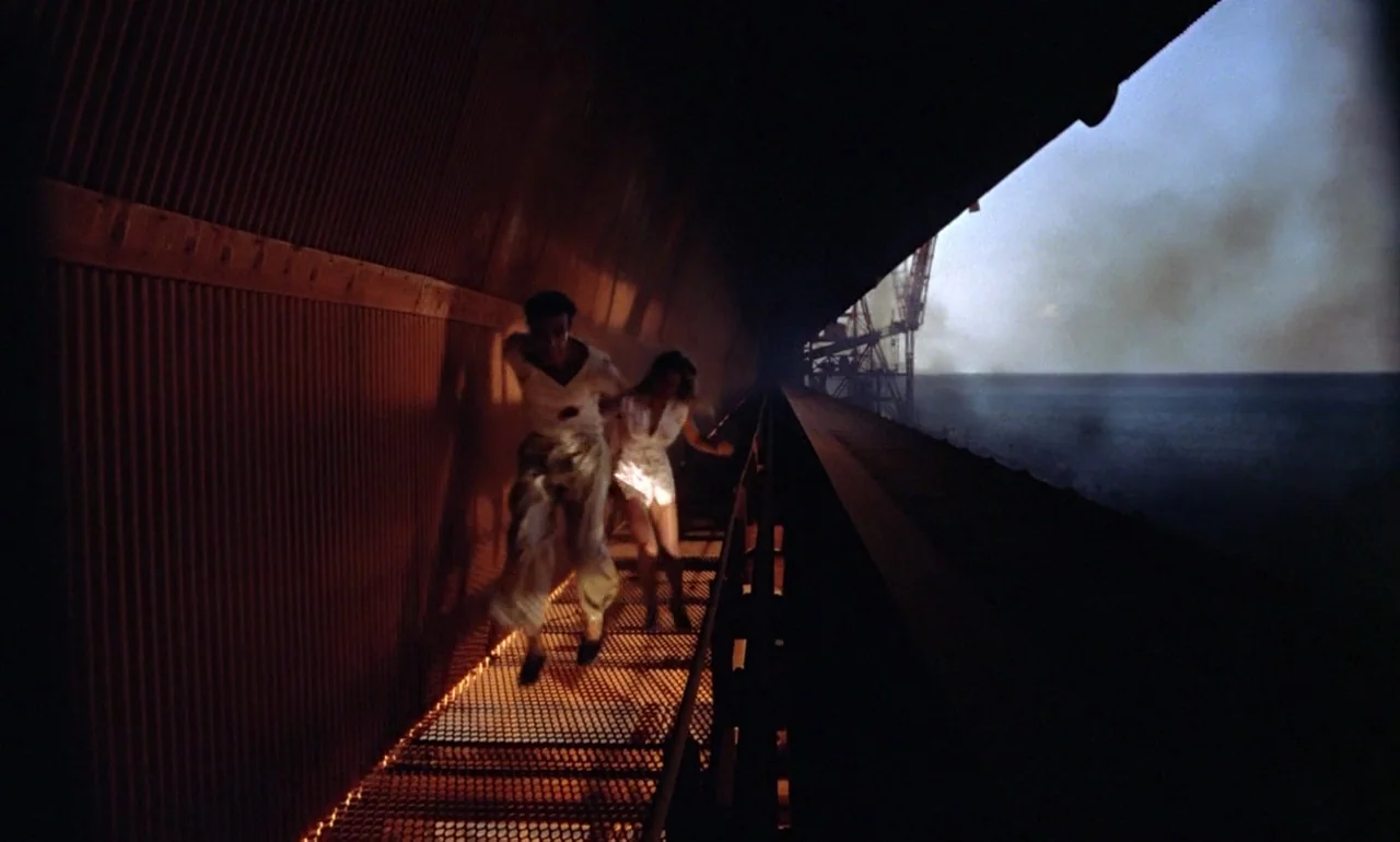 Dr. No (1962) movie still: fullbody — A dark, corrugated metal structure borders a narrow grated walkway above the ocean. Two people run forward…