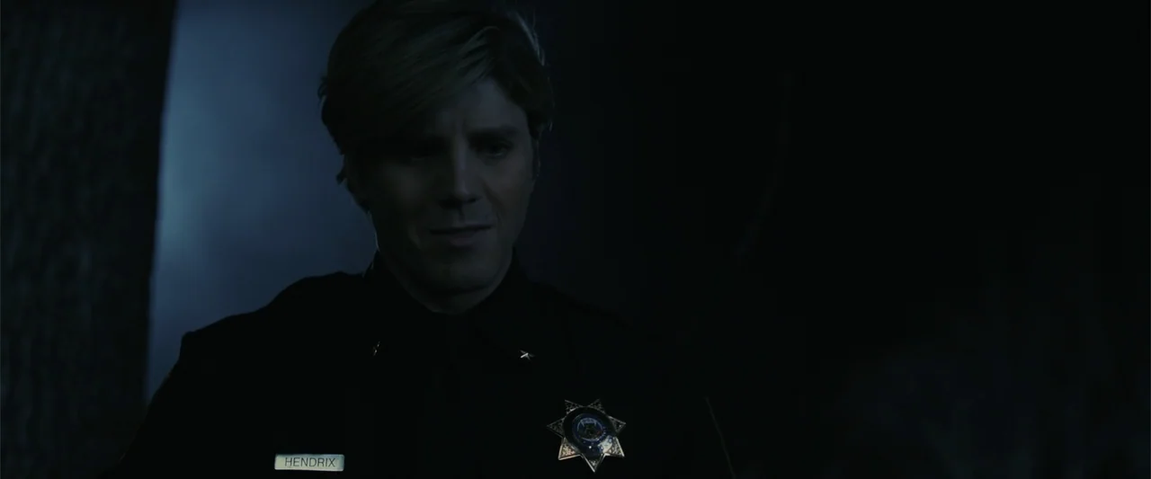 Companion (2025) movie still: medium — A dark, wooded area provides a backdrop for a police officer at night. The officer in a uniform stares ahead…