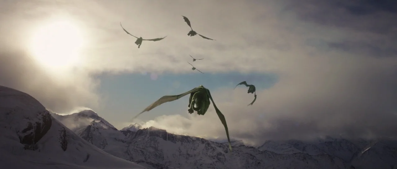 Pete’s Dragon (2016) movie still: wide — A vast snow-covered mountain range sits under a cloudy, bright sky. Several winged reptiles fly in a…