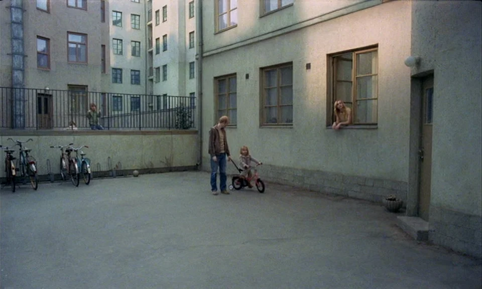 You, The Living (2007) movie still: wide — A quiet apartment courtyard with light-colored stucco buildings surrounds a paved concrete area. A man…