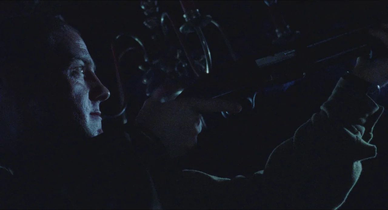 Dog Soldiers (2002) movie still: medium — A dark indoor space contains a man aiming a firearm towards the right. Behind him stands an ornate metal…