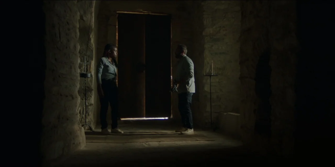 The Hunted (2024) movie still: fullbody — A stone corridor with rough walls is illuminated by bright light from a doorway. Two people face each…