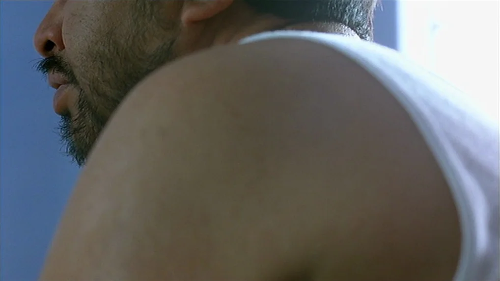 Battle In Heaven (2005) movie still: closeup — A man in a white undershirt sits against a plain blue wall. The man is positioned in side profile…
