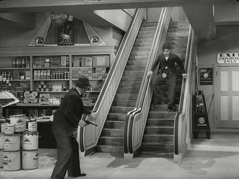 Modern Times (1936) movie still: fullbody — A department store interior features shelves with retail products and a moving escalator. A man in a…